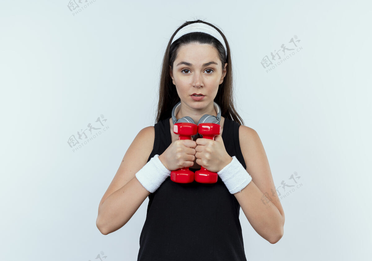 站立戴着头巾的年轻健身女 正脸举着哑铃站在白墙上锻炼健身年轻女人