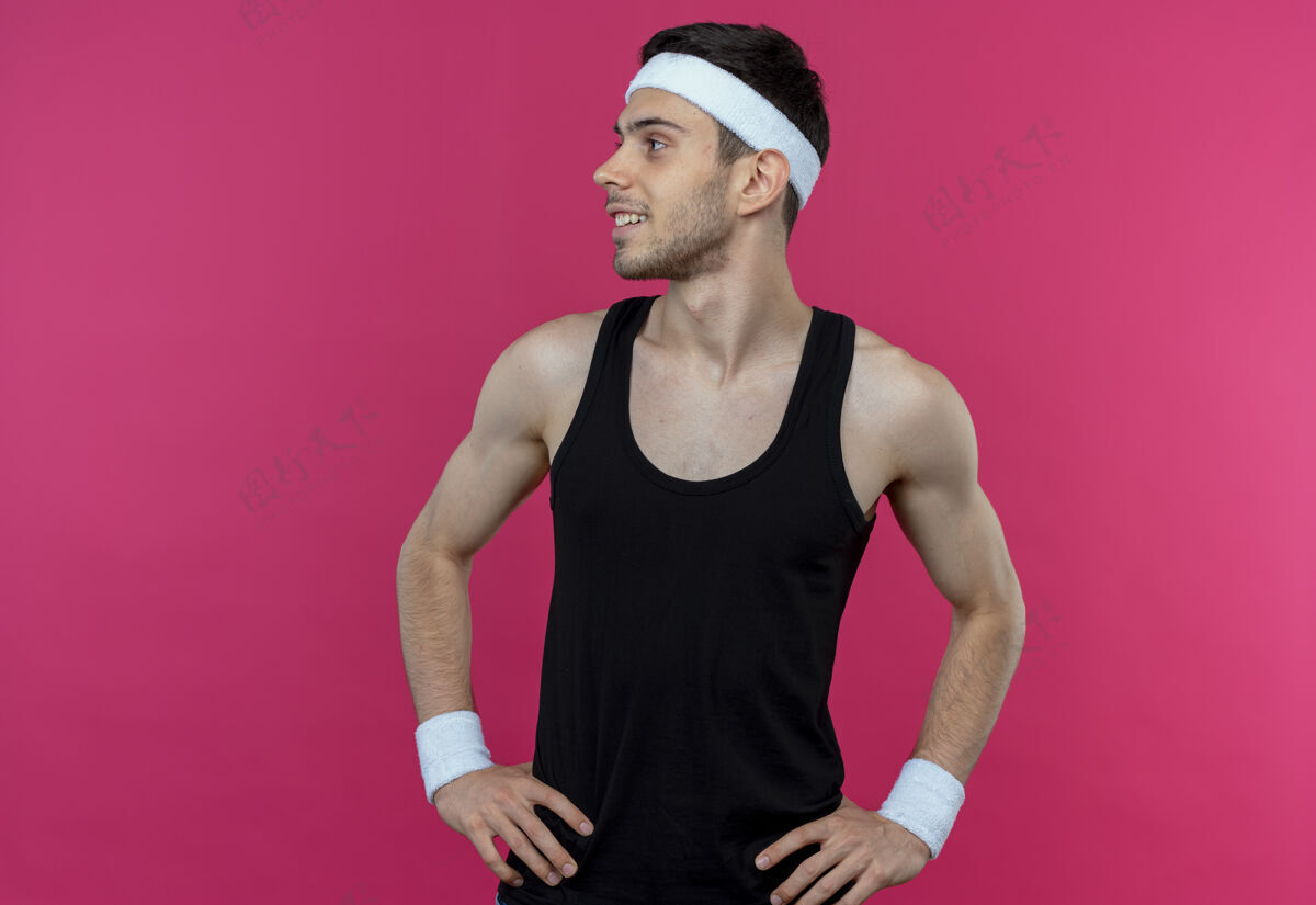 年轻戴着头巾的年轻运动型男子站在粉红色的墙上 两臂叉腰 微笑着向一边望去站着头带臀部