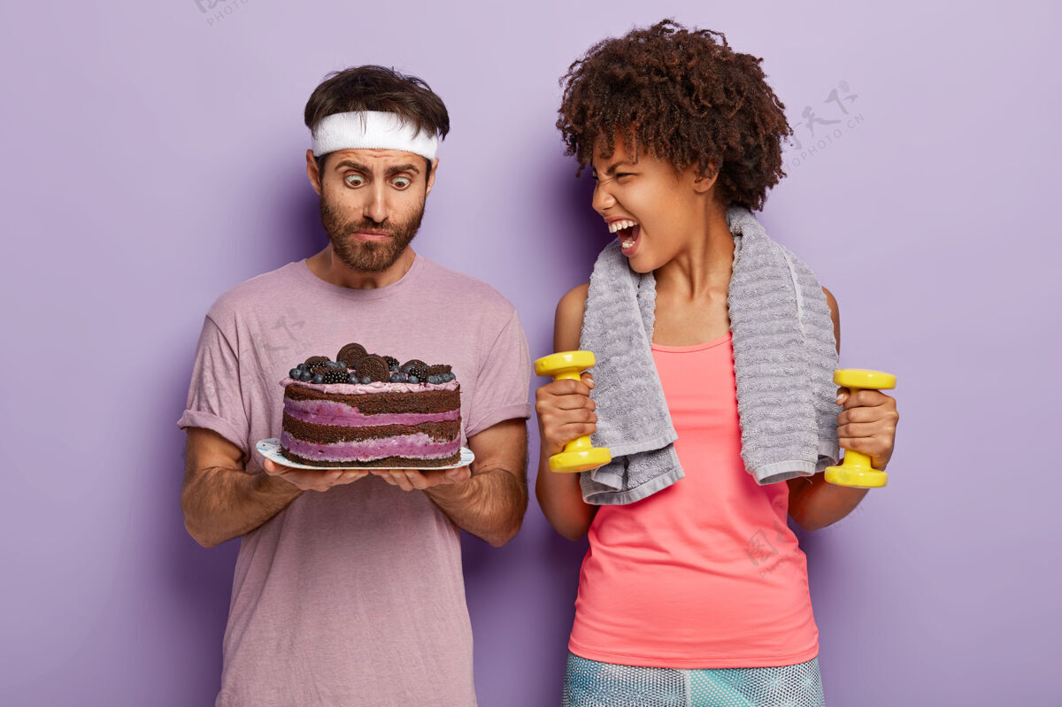 男惊讶的男人盯着烤好的甜饼 感觉到诱惑 情绪激动的女人对着他大叫 手里拿着黄色的哑铃女士情侣一起