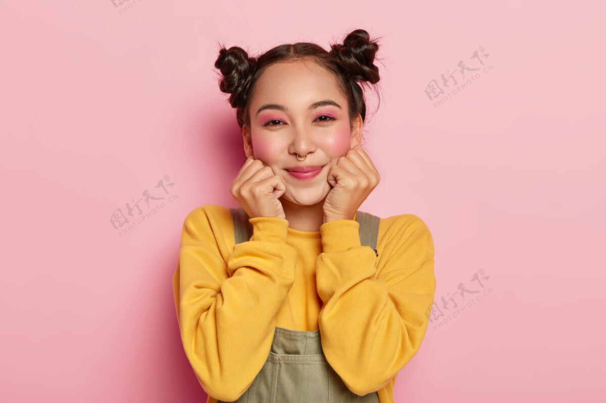 肖像快乐的亚洲女孩的画像 浓妆艳抹 深色头发梳成两个发髻 鼻子穿孔 穿着休闲的黄色运动衫和工作服套装微笑女人