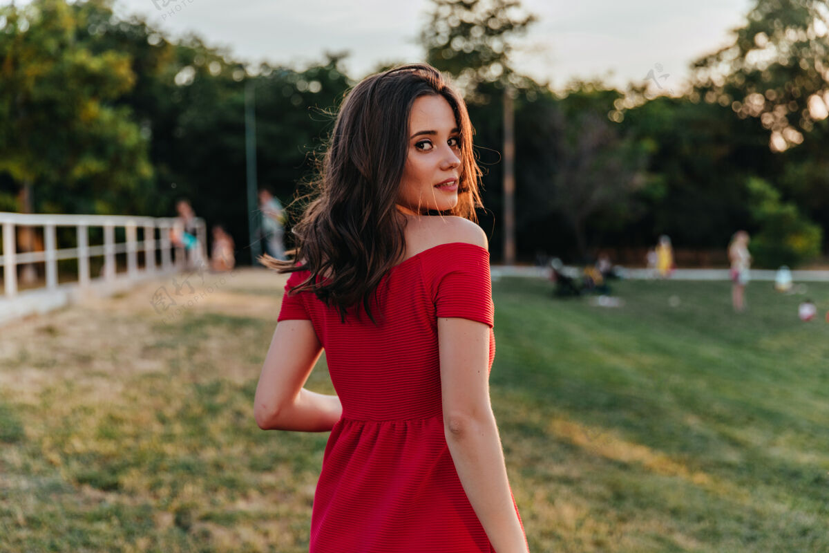 女人梦幻般的棕色头发的女孩在公园里散步时回头看穿着红色裙子站着的迷人的高加索女士的肖像草快乐美丽
