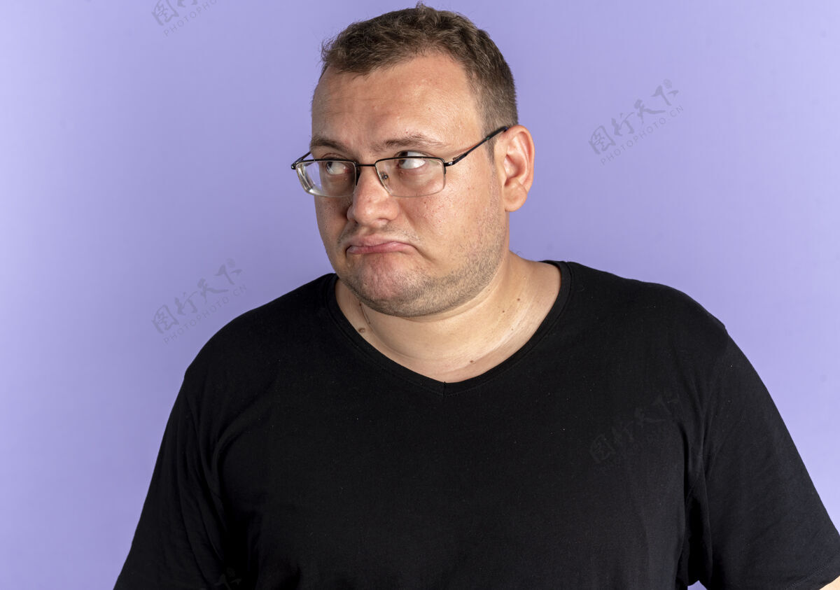 男人一个戴着眼镜 身穿黑色t恤的超重男子站在蓝色的墙上 表情悲伤地看着一边表情表情超重