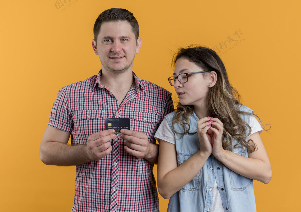 手机一对幸福的年轻夫妇 男人和女人在使用电子产品 女人低头看着她男友的智能手机屏幕 站在橙色的墙上男朋友向下小玩意
