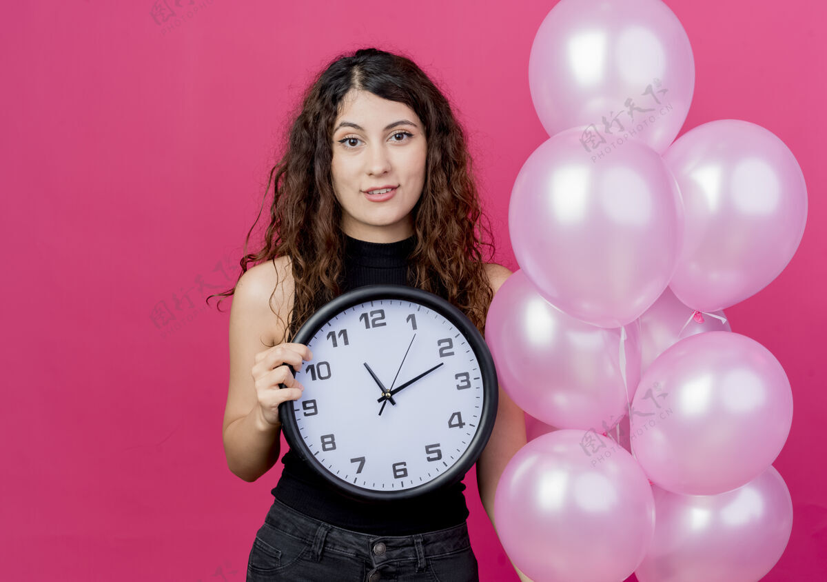 微笑一位年轻漂亮的卷发女士手持一束气球 站在粉红色的墙上高高兴兴地微笑着欢呼时钟空气
