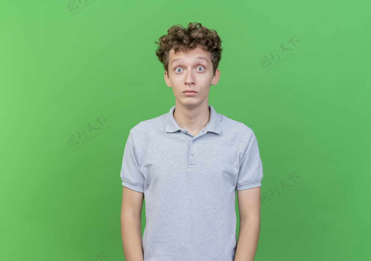 看穿着灰色马球衫的年轻人站在绿色的墙上睁大了眼睛 既惊讶又惊奇惊人站年轻