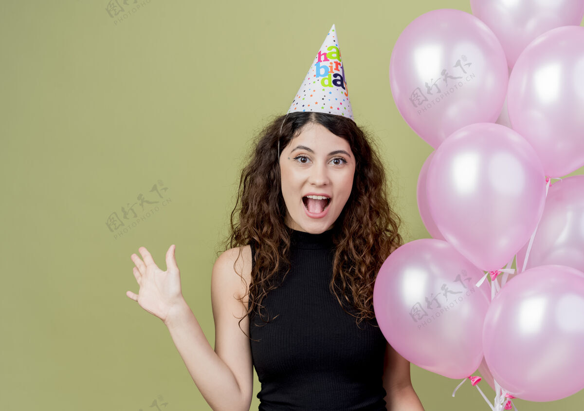 空气一位年轻漂亮的女士 卷发 戴着节日帽 手持气球 举手 高兴而兴奋地微笑着 生日派对的概念站在光墙上年轻微笑卷曲