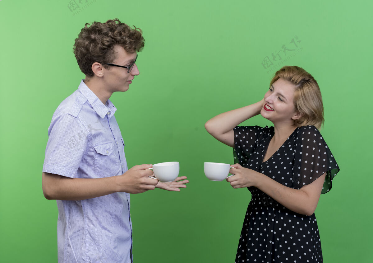积极年轻漂亮的夫妇拿着咖啡杯面对面站着 微笑着 幸福而积极地站在绿色的墙上站着杯子夫妻