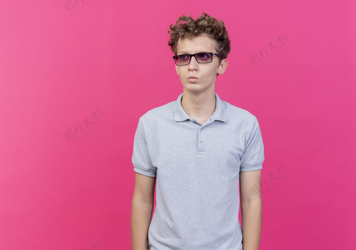 脸一个戴着黑眼镜 穿着灰色马球衫的年轻人站在粉红色的墙上 严肃地看着一边戴年轻站