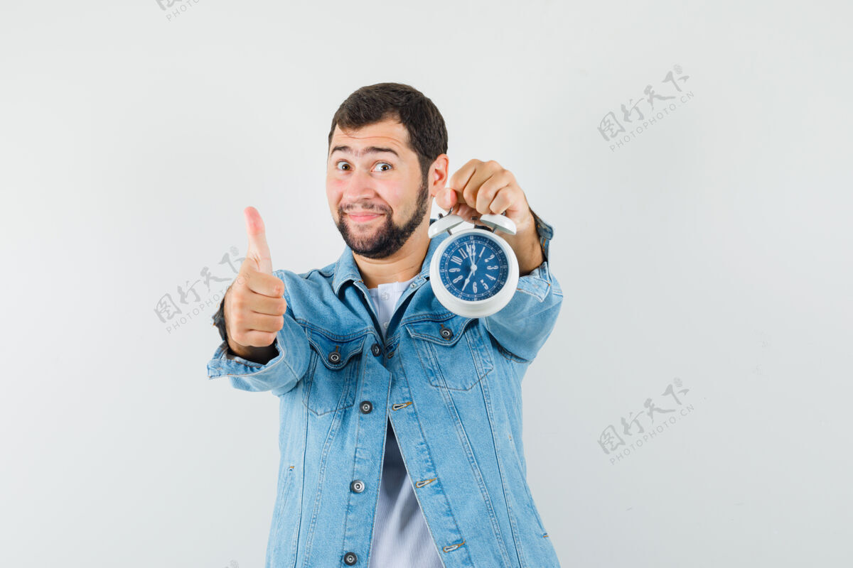 经典复古风格的男士夹克衫 t恤展示拇指向上 同时手持时钟 看起来很满意 正面视图显示拇指年轻