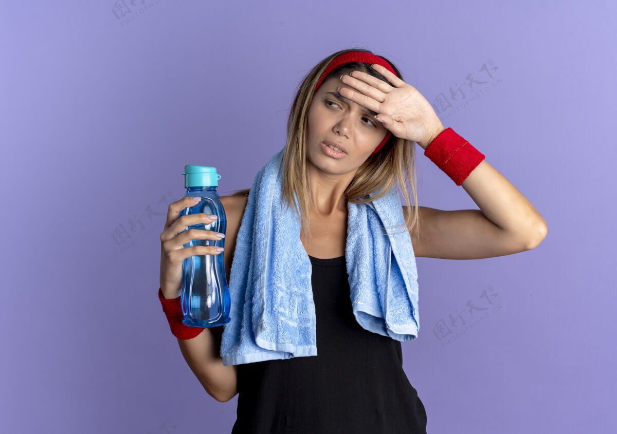 疲惫穿着黑色运动服 头戴红色头巾 脖子上围着毛巾 手里拿着一瓶水的年轻健身女孩站在蓝色的墙壁上 看起来很疲惫 筋疲力尽毛巾奶瓶年轻