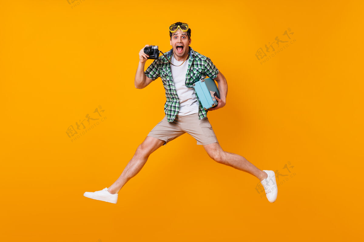 行李一个穿着夏季服装的有趣的游客 手里拿着复古相机和蓝色手提箱一个戴着潜水面具的人在橙色的空间里跳跃使机票休闲