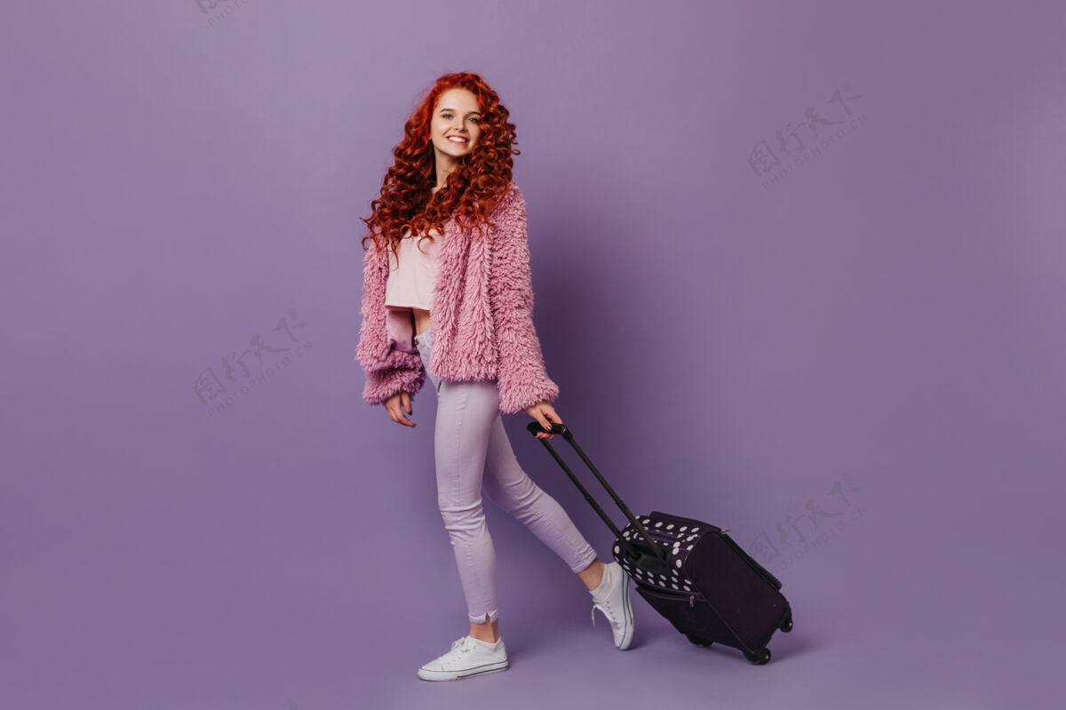性感积极的蓝眼睛女孩 红色卷发 穿着时髦的皮草外套和白色裤子 提着手提箱手提箱少女粉色外套