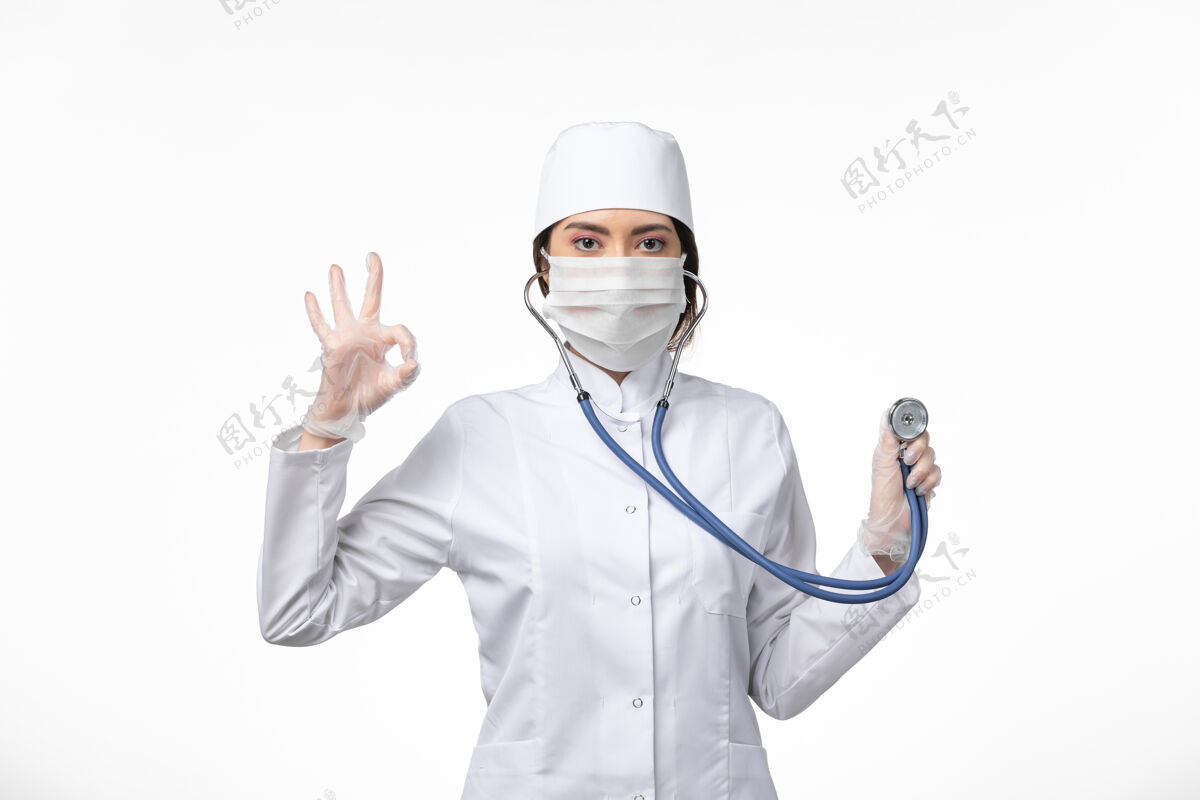 专业人员正面图女医生身穿白色无菌医疗服 戴口罩因冠状病毒-使用听诊器对白墙疾病冠状病毒-大流行性疾病冠状病毒口罩听诊器