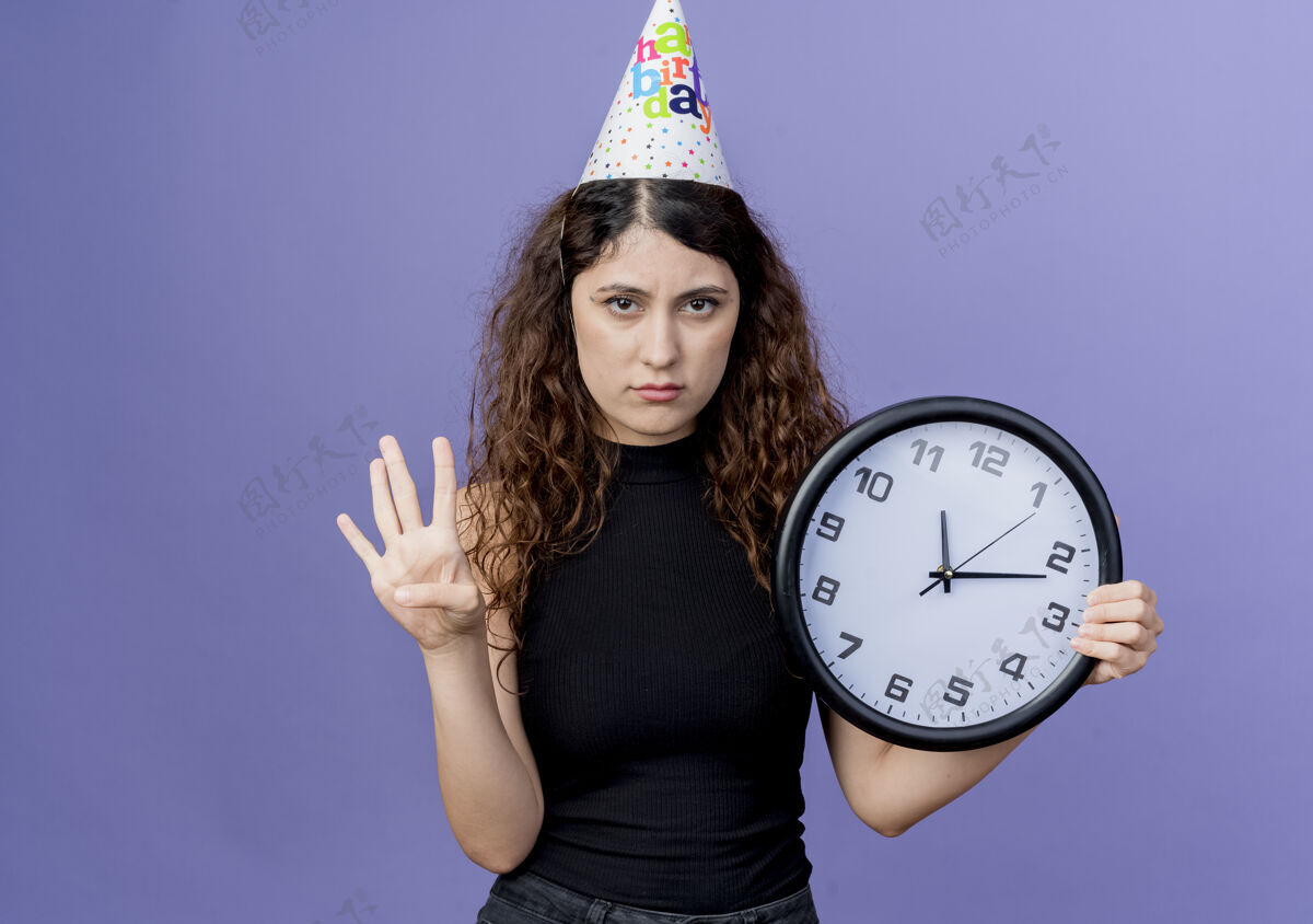 悲伤一个年轻漂亮的女人 卷发 戴着节日帽 手里拿着挂钟 上面写着第四个悲伤的表情生日派对的概念站在蓝色的墙上表情抱着钟