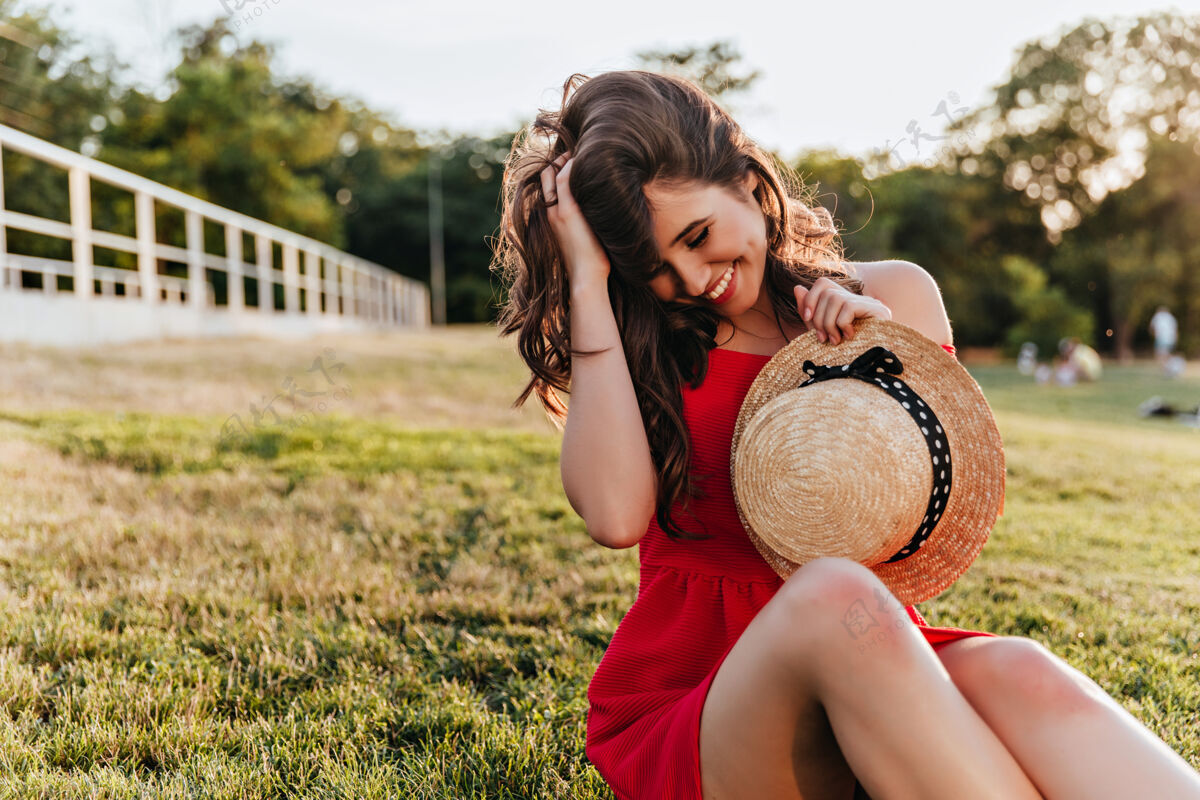 女人情绪化的黑发女孩戴着优雅的帽子在公园里休息穿着红色衣服的迷人女士坐在草地上抚摸着她的头发草优雅公园