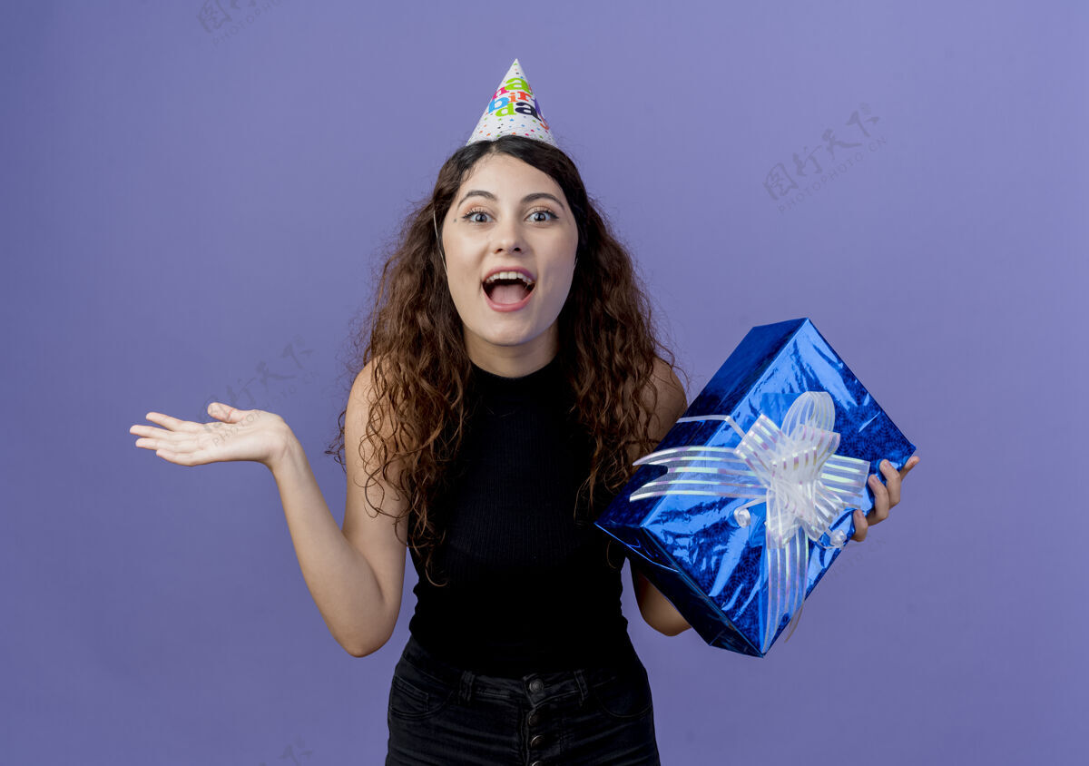 微笑一个年轻漂亮的女人 卷发 戴着节日帽 手里拿着生日礼盒 脸上带着困惑和惊讶 脸上洋溢着快乐的笑容 站在蓝色的墙上庆祝生日惊喜卷发站着