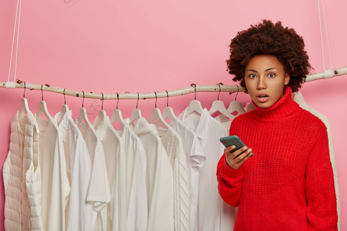室内尴尬的美国黑人妇女用手机购物 在服装店摆姿势 穿红色毛衣 在衣架旁摆白色衣服黑色震惊一