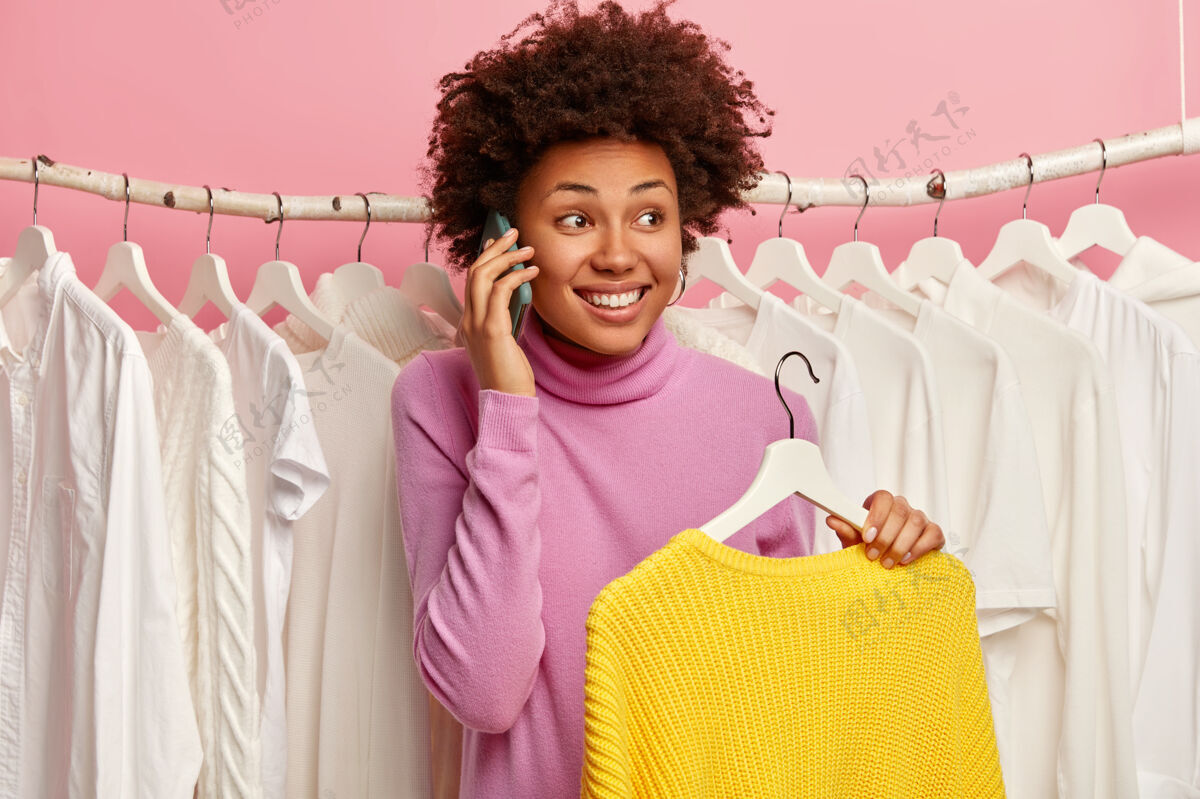 粉色喜出望外的女人打电话 站在满是白色衣服的衣架旁 拿着针织的冬季黄色毛衣 享受在时尚商场购物的日子购买非洲购买