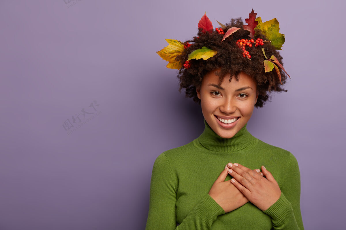 手臂可爱的年轻美国黑人女士感激不尽 双手合十放在胸前 绿色长袖套头衫 卷发上的叶子快乐表情叶子