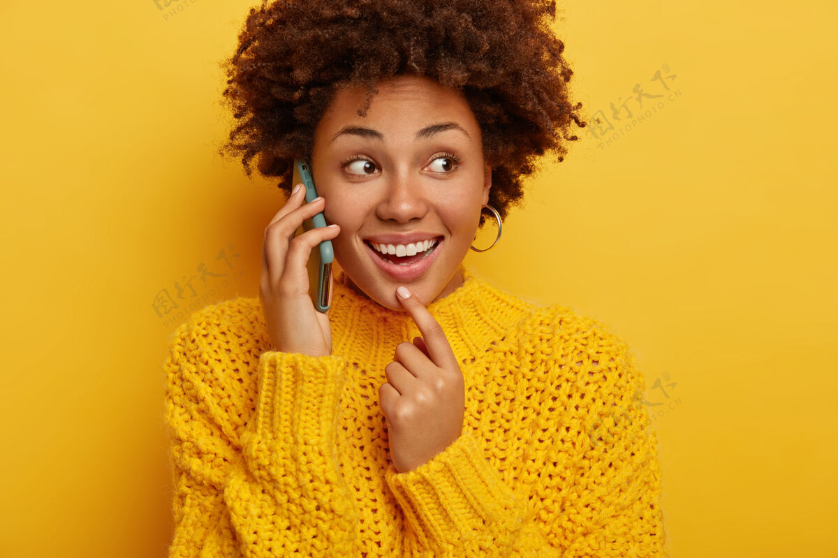 手机一个快乐的非洲裔美国女人的剪短镜头有电话交谈 把小玩意儿放在耳边 注意力集中在一边 穿着暖和的衣服 隔离在黄色背景下休闲微笑服装