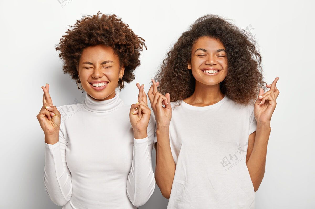 青年两位充满希望的美国黑人妇女祈祷 双手交叉 笑容灿烂 身着白色休闲服 期待积极的结果 并肩站在一起梦想朋友牙齿