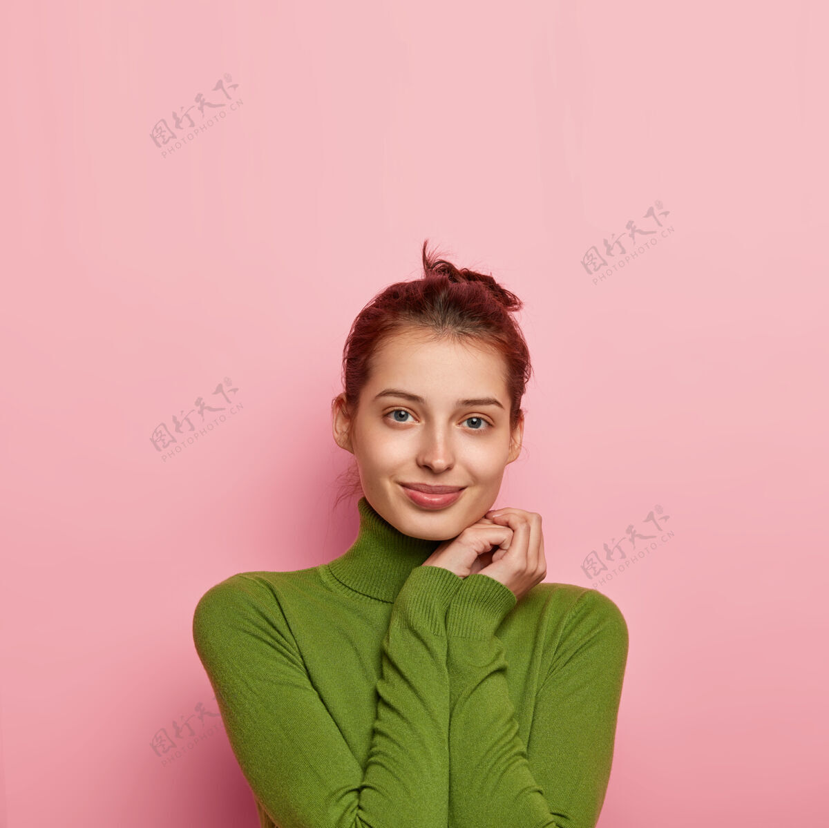 发髻迷人的欧洲女人的垂直镜头保持双手靠近脸部 穿着休闲的绿色高领毛衣 直视镜头 隔离在粉色背景上 为您的自由空间满意眼睛年轻