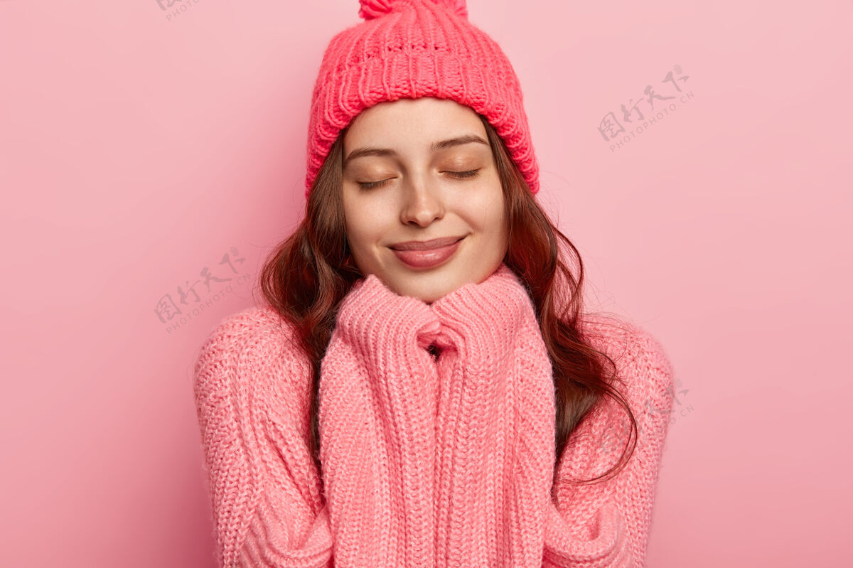 罗西满意的欧洲女人的头像有健康的皮肤 闭着眼睛 手放在下巴下 戴着暖和的帽子和超大的毛衣 隔离在粉红色的背景上温暖健康满意
