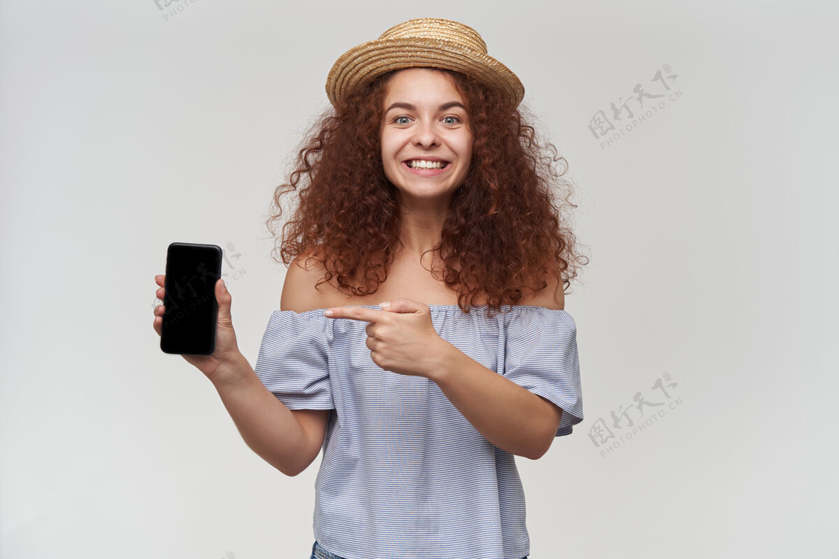 女性漂亮的女人 姜黄色卷发的女孩穿着条纹露肩上衣和帽子拿着和指着智能手机 复制空间隔离在白色的墙壁上面部室内青少年