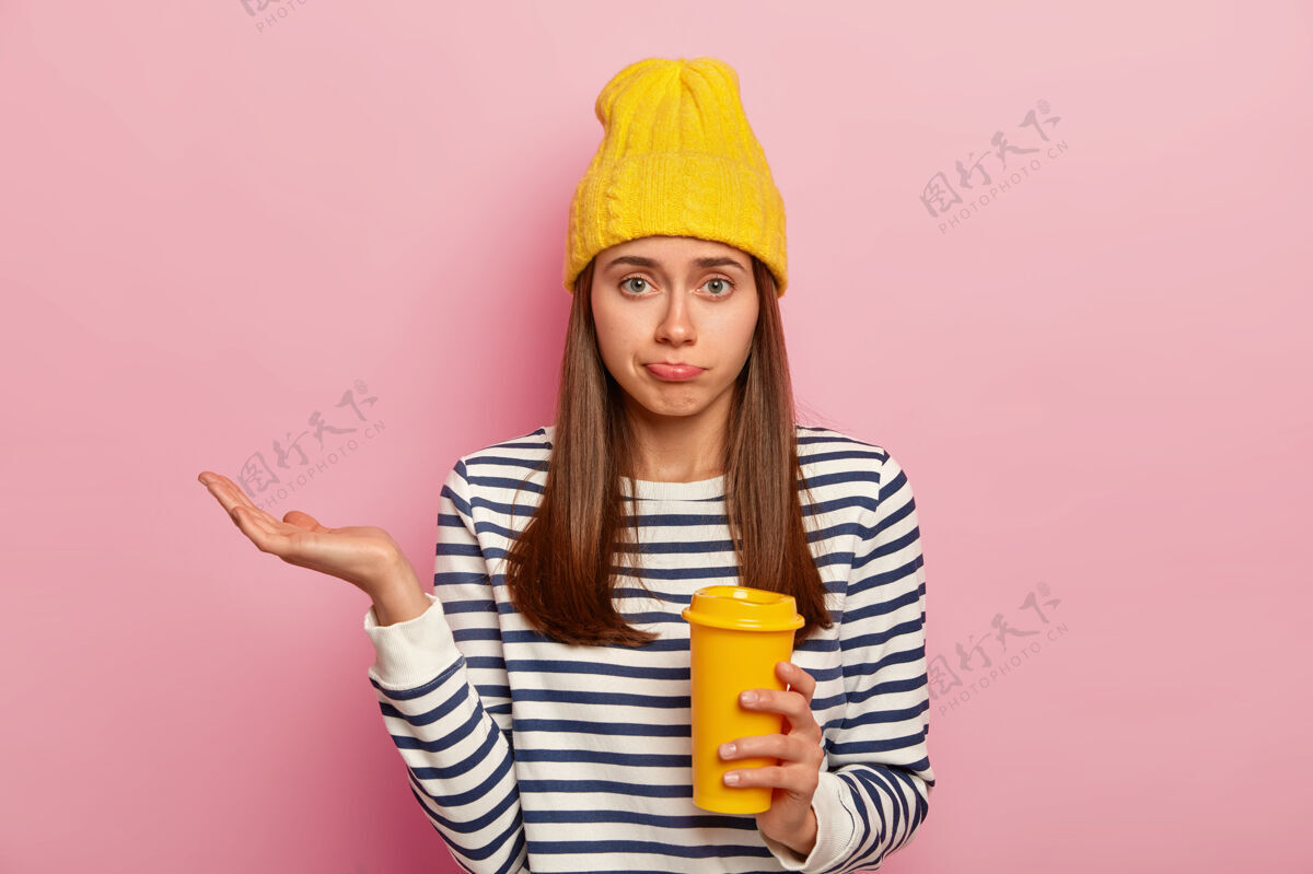 白种人犹豫不决的欧洲女人的照片 表情混乱 举起手掌 拿着外卖咖啡 戴着黄色帽子和条纹毛衣 在粉色背景上摆姿势决定拼图室内