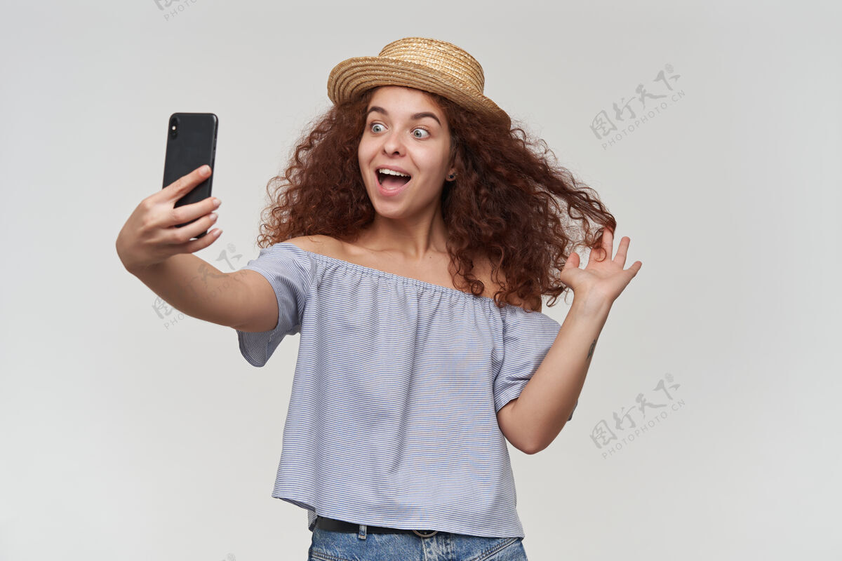 红发有魅力的红头发卷发女孩的肖像穿着条纹露肩衬衫和帽子在智能手机上自拍 玩头发和微笑孤立地站在白墙上姜黄色心情微笑