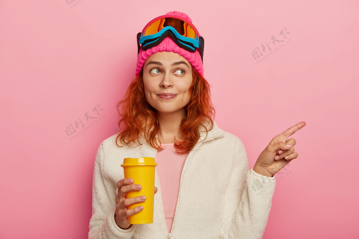 帽子美丽的红发女孩的半身镜头指向右边 向度假地的游客指明方向 有活跃的冬季休息 喝外卖咖啡 戴着滑雪面具 隔离在粉色背景上滑雪板高兴路