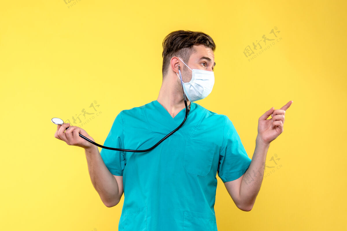 健康黄色墙上带听诊器和面罩的男医生的正面图男医生听诊器微笑