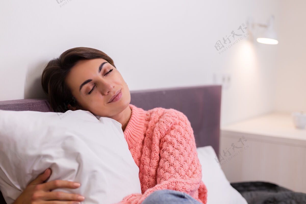 快乐穿着粉色可爱针织套头衫的美女躺在家里的床上 微笑着 享受独处的时光房间时尚女人