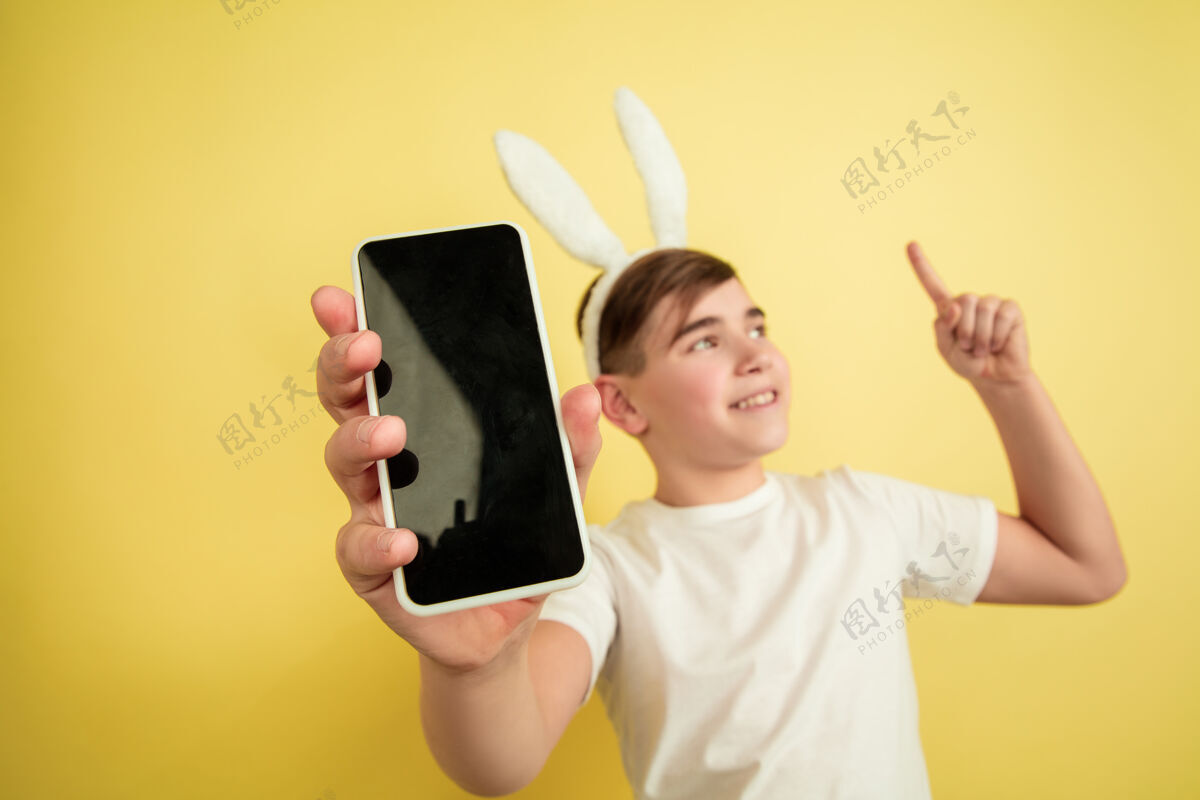 复制空间显示空白屏幕 指向上白人男孩作为复活节兔子在黄色背景上复活节快乐问候有趣五颜六色表情