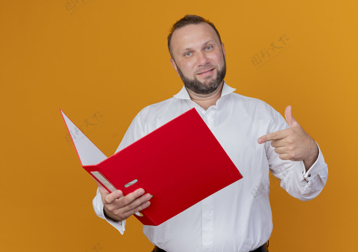 握一个留着胡子的男人穿着白衬衫 拿着文件夹 用手指着它 微笑着站在橙色的墙上笑站手指