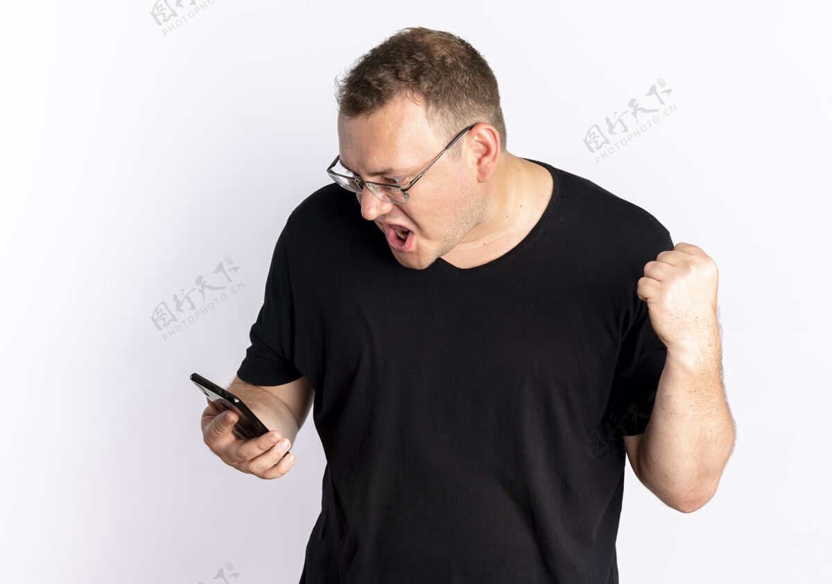 站一个戴眼镜 身穿黑色t恤的超重男子站在白墙上看着智能手机屏幕 握紧拳头 愤怒的脸脸男人手机