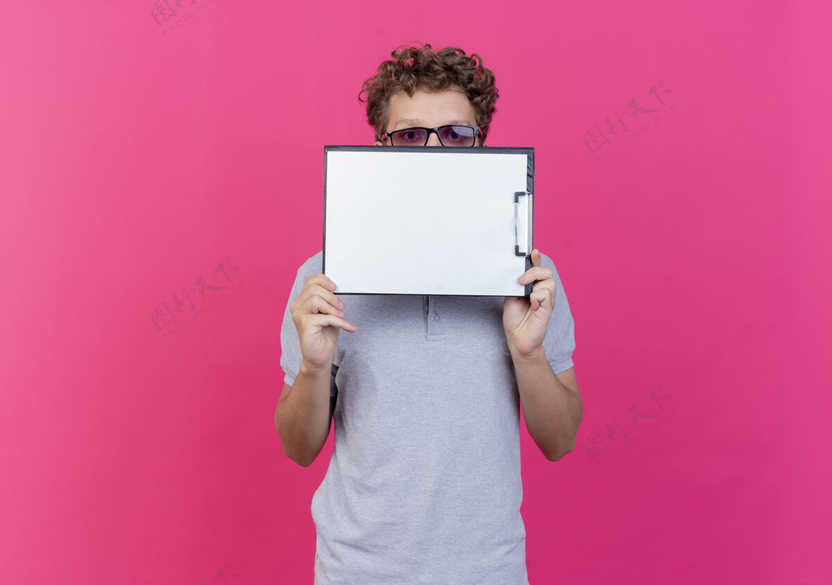 眼镜一个戴着黑眼镜 穿着灰色马球衫的年轻人拿着一张白纸夹在剪贴板后面 站在粉色的墙上偷看马球男人立场