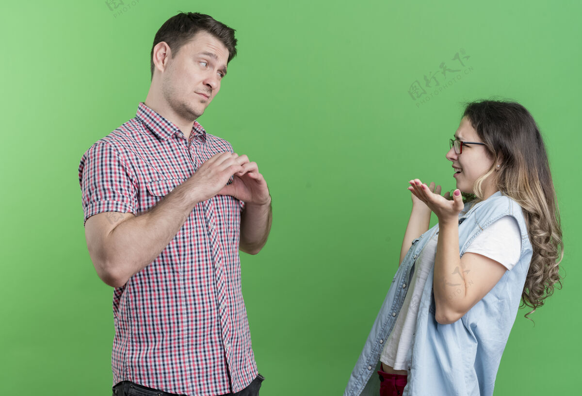 心一对年轻夫妇穿着休闲服的男人和女人伤心的男人向站在绿色墙壁上的她不高兴的女友展示心形情侣显示姿态