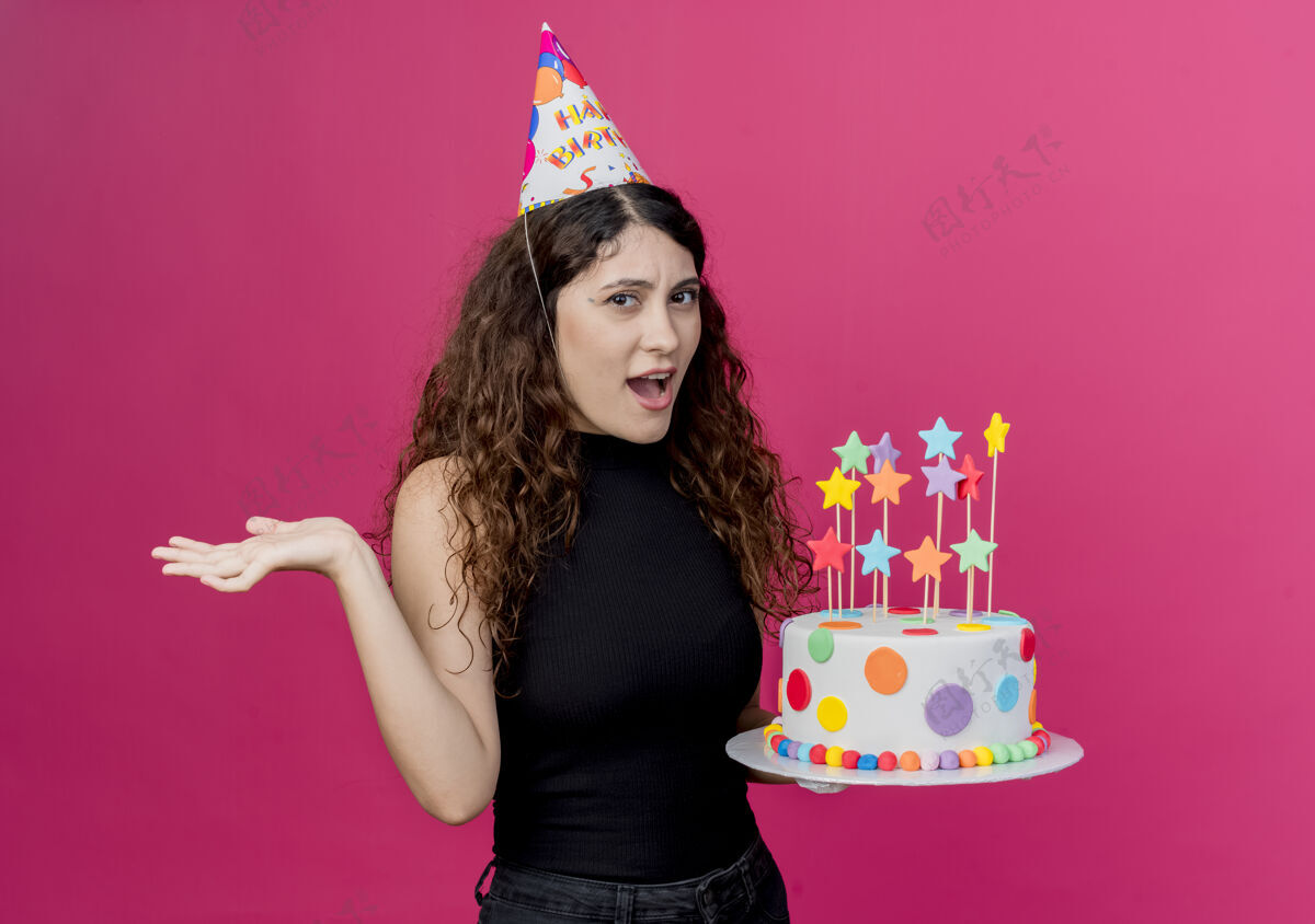 帽子一个年轻漂亮的女人 卷发 戴着节日帽 手里拿着生日蛋糕 站在粉色的墙上 困惑着生日派对的概念年轻卷发困惑