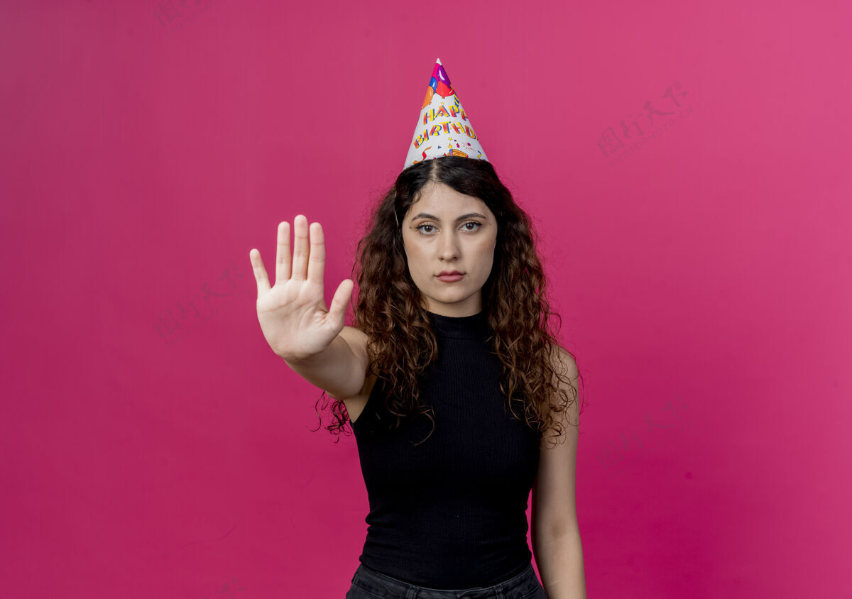 脸一位年轻漂亮的女士 卷发 戴着节日帽 严肃的表情 站在粉色的墙上写着生日派对的标志站看严重