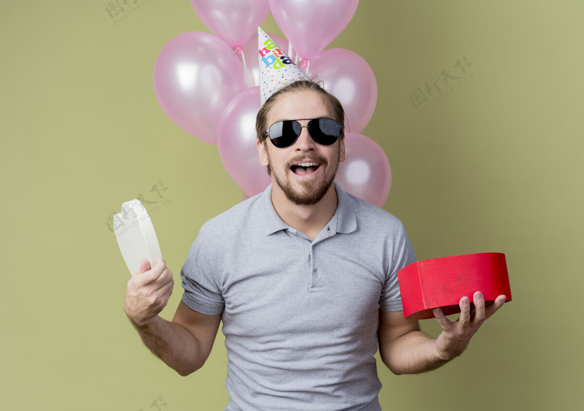 举行戴着节日帽的年轻人举着礼盒庆祝生日快乐而兴奋地微笑着 手里拿着气球站在轻墙上气球光年轻