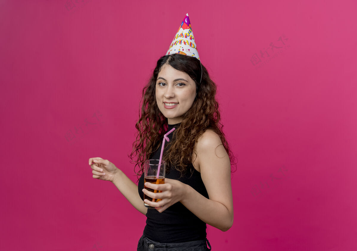 卷发一位年轻漂亮的女士 一头卷发 戴着节日帽 手里拿着鸡尾酒 微笑着 站在粉色的墙上微笑年轻帽子