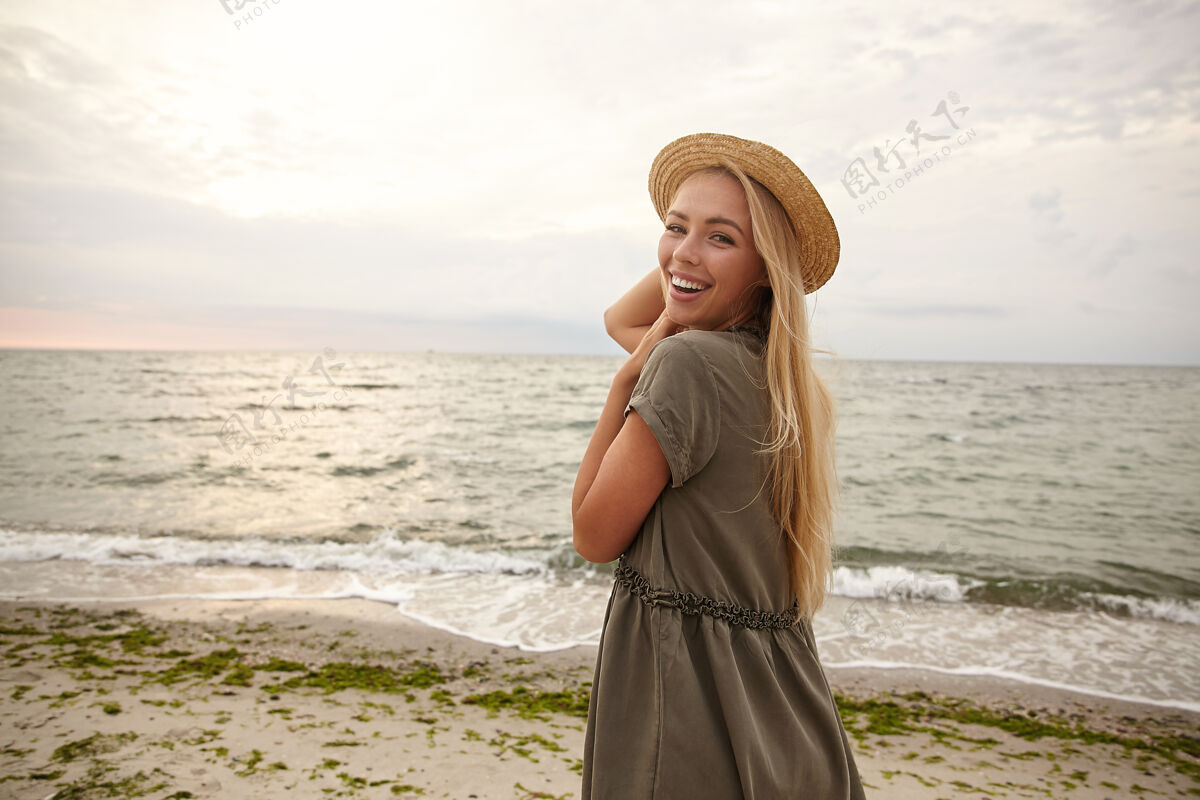 白皙户外拍摄的快乐的年轻迷人的长发金发女子保持她的帽子与举起的手 笑着高兴地看着她的肩膀减肥年轻欢呼