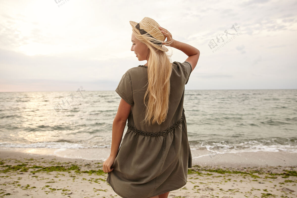 波浪水平拍摄年轻迷人的长发金发女子不断提高她的手在船上的帽子 并微笑着高兴地在海边摆姿势与她的背部海边女人长