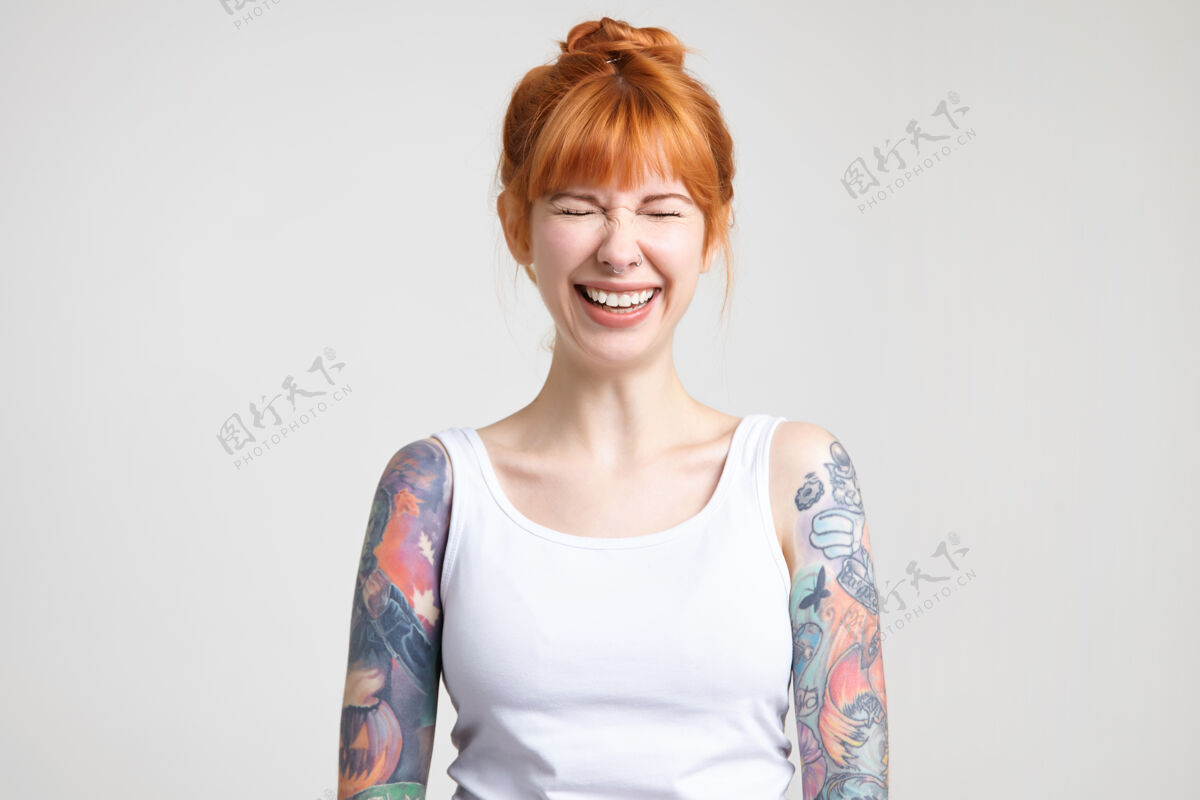 姿势快乐的年轻迷人的红发女子纹身保持她的眼睛闭上 而欢笑 穿着白衬衫 而在白色的背景摆姿势塔托鼻子衬衫