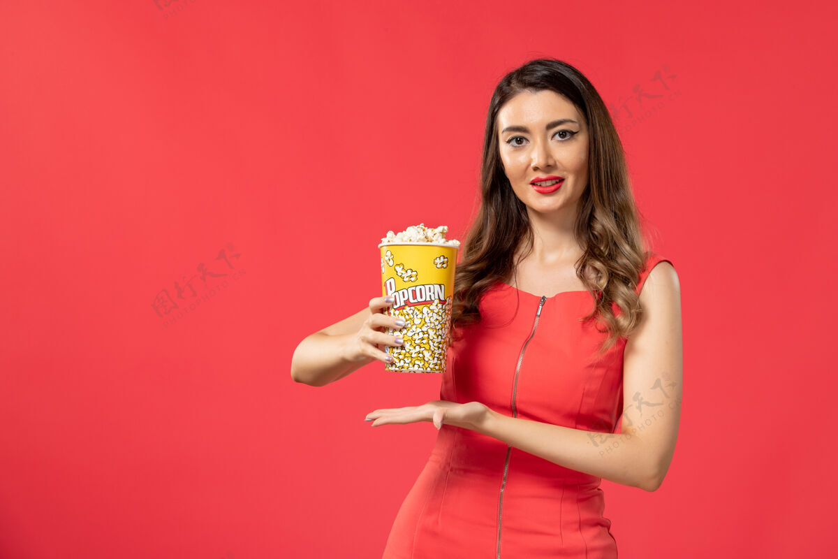 电影正面图身穿红色衬衫的年轻女性手持爆米花包 在红色表面微笑漂亮女士包装