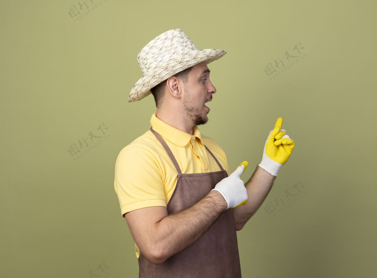 帽子年轻的园丁穿着连体衣戴着帽子戴着工作手套用食指指着背看着困惑看站花园