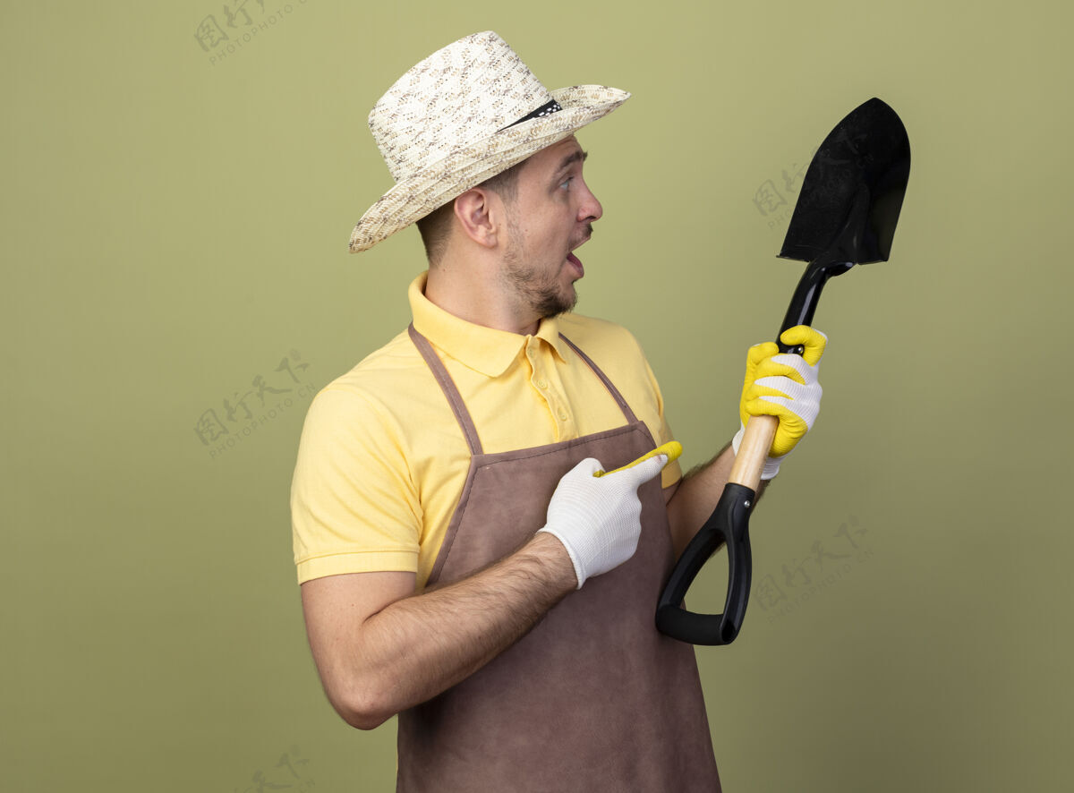 目录年轻的园丁穿着连体衣戴着帽子戴着工作手套拿着铲子用食指指着它 看上去很惊讶年轻人看手套