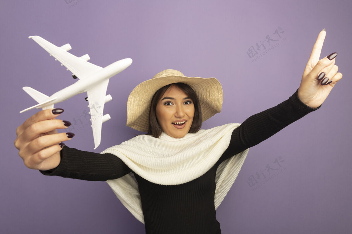 围巾戴着白色围巾 戴着夏帽的年轻女子展示着玩具飞机快乐欢快的微笑 食指朝上手指看飞机