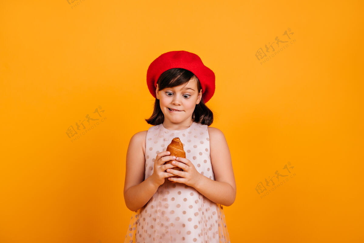 法国抱着羊角面包的小女孩戴着法国贝雷帽的黑发小孩在黄色的墙上表示惊讶羊角面包微笑食物